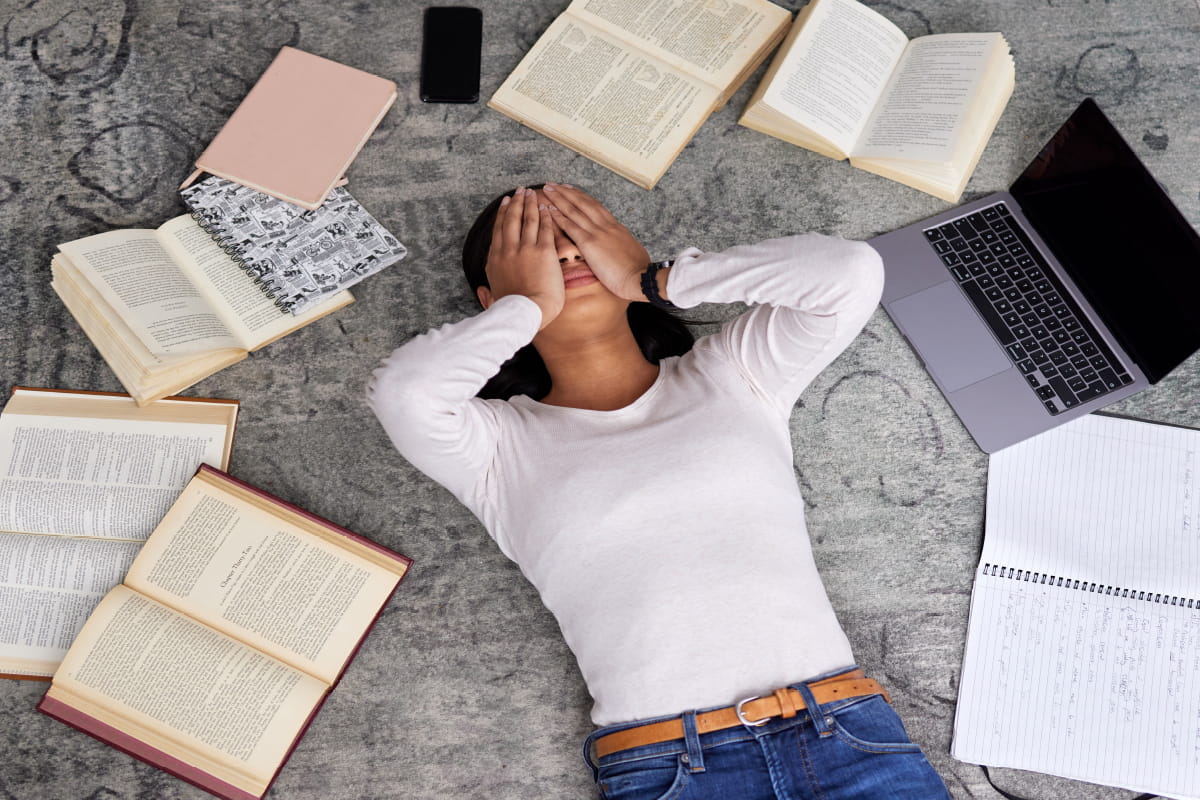Student laying on the floor with books open all around them with their hands covering their face.