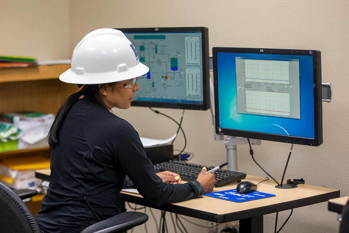 An individual in a white hard hat and safety goggles sits at a desk looking at a computer screen