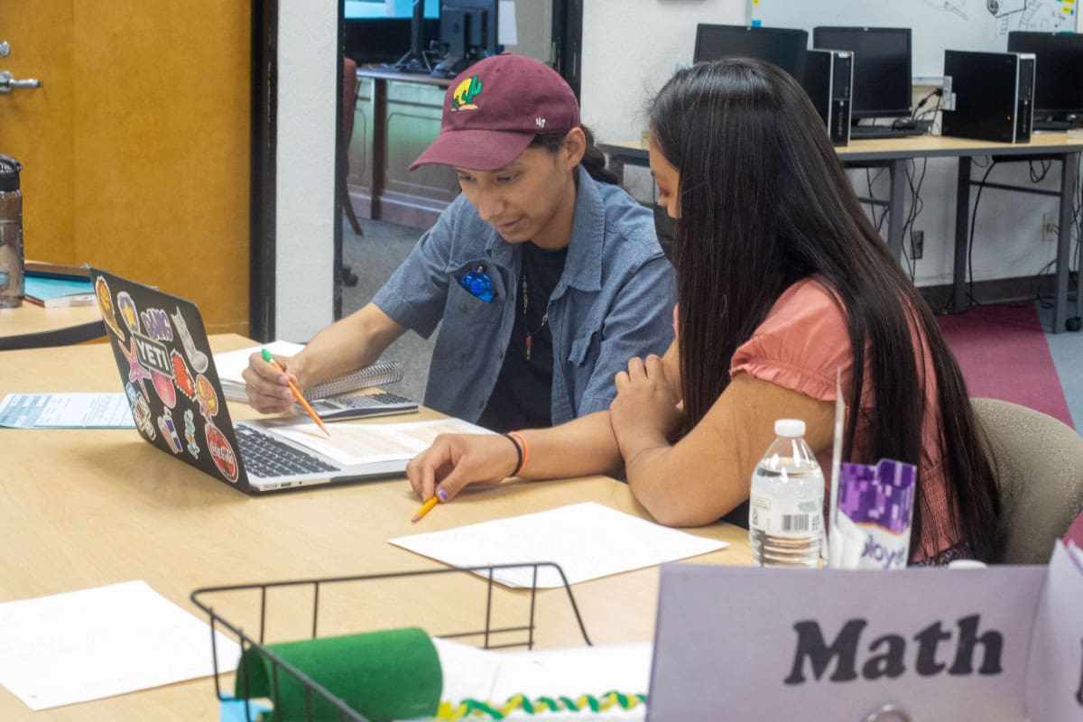 Two individuals sit at a table with an open laptop in front of them, reviewing text on a piece of paper