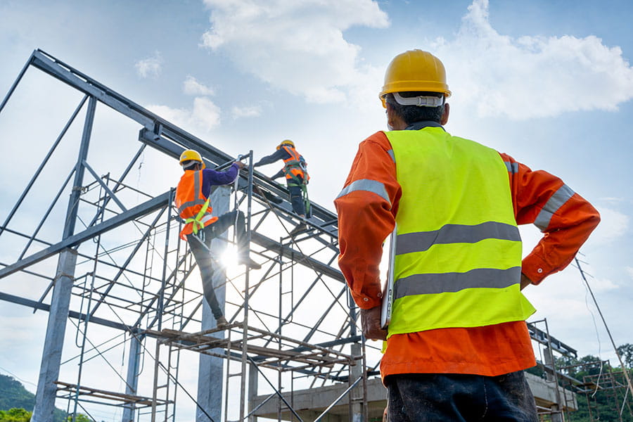 An individual in a yellow safety vest and hard hat watches two other individuals in similar attire climbing scaffolding