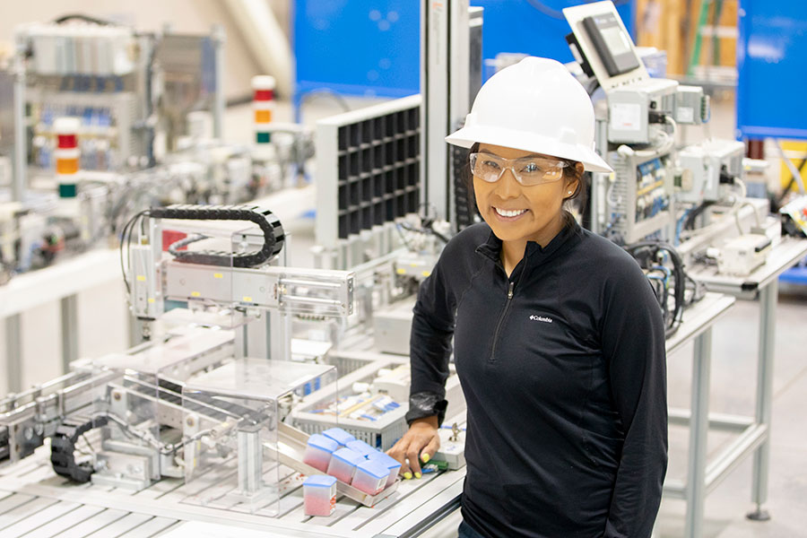 An individual wearing a white hard hat and safety goggles smiles while standing in front of machinery