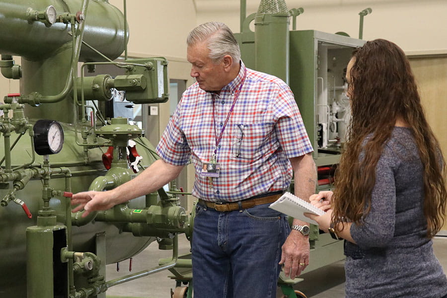 An individual touches valves on machinery while another individual looks on and takes notes in a notebook