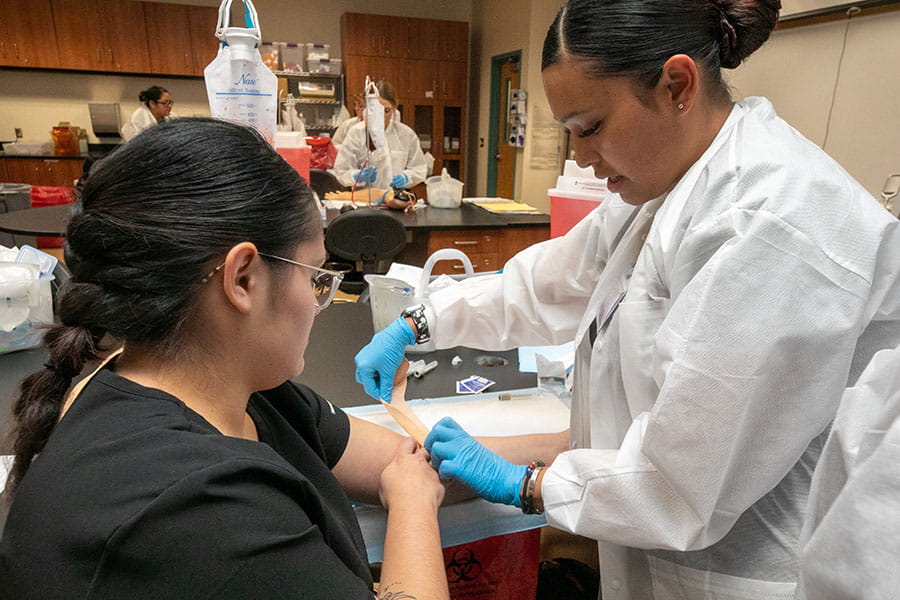 A student in lab coat demonstrates applying a bandage wrap on another student