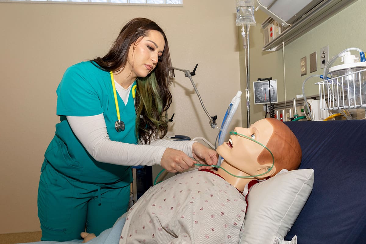 Student wearing scrubs adjusts equipment on training manikin.