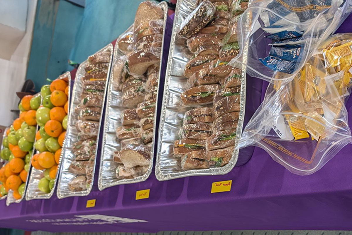 A purple table with variety of sauce packets, trays of sandwiches, and trays of oranges and green apples.